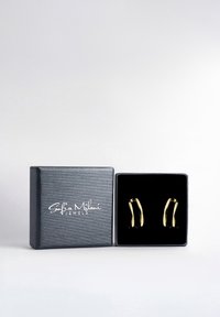 Guldörhängen i form av hoops visade i en svart sammetklädd låda med en texturerad mörk utsida och en vit logotyp på locket.