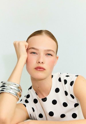 Jeune femme aux cheveux lissés en arrière reposant sa tête sur son poing gauche, vêtue d'un haut blanc sans manches à pois noirs et de bracelets argentés.