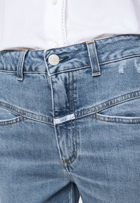 Jean en denim bleu clair avec un design classique à cinq poches, doté d'une fermeture à bouton en métal, de coutures jaunes et de détails légèrement déchirés.
