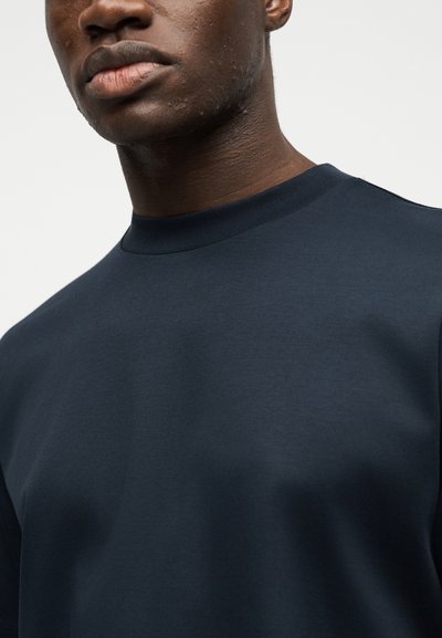 Κοντινό πλάνο ατόμου που φοράει απλό σκούρο μπλε μπλουζάκι με στρογγυλή λαιμόκοψη, εμφανίζοντας το κατώτερο μέρος του προσώπου και το πάνω μέρος του κορμού με φόντο ανοιχτόχρωμο.