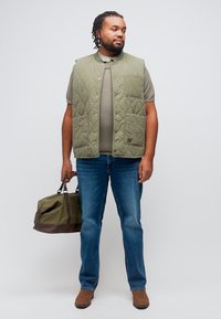 Άνδρας με πλεξούδες φοράει πράσινο καπιτονέ γιλέκο, γκρι πουκάμισο, μπλε τζιν, καφέ παπούτσια, κρατώντας πράσινη και καφέ τσάντα duffel, στεκόμενος μπροστά σε λευκό φόντο.