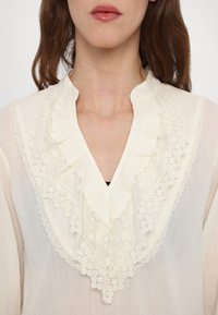 Le haut du torse d'une femme portant un chemisier crème avec un col en V à volants et des détails en dentelle florale le long du col et de la poitrine.