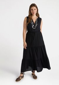 Abito maxi senza maniche di colore nero con gonna a strati, vita con cordino e collana decorativa. Indossato con espadrillas nere in rete.