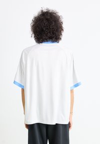 Camisola desportiva branca com mangas curtas, apresentando um colarinho e acabamentos de mangas em azul claro sólido. Textura suave, corte relaxado e design minimalista.