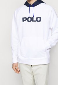 Sudadera blanca con capucha y detalles azules, que presenta un gran logotipo negro de "POLO". Tejido suave con un bolsillo delantero y puños acanalados.