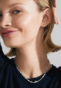 Sidabrinė grandinėlė su gėlių akcentais ir perlais, vilkima prieš tamsų tekstūruotą megztinį. Artimas veido vaizdas, plaukai iš dalies surišti.