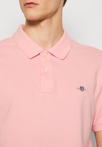 Muž nosi svetloružové tričko polo s krátkym rukávom, dvoma gombíkmi a malým vyšívaným logom GANT na ľavej strane hrudníka.