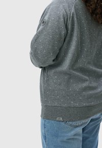 Felpa grigia con superficie texturizzata, caratterizzata da un sottile motivo a pois, polsini a costine e un piccolo etichetta con logo sull'orlo. Abbinata a jeans blu.