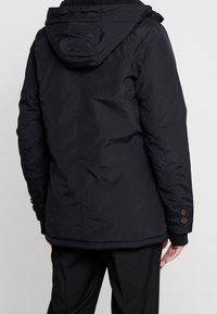 Svart isolerad jacka med huva, texturerad tyg och knappdetaljer på ärmarna. Snygg, förlängd design, lämplig för kallt väder.