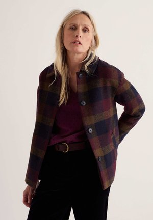 Femme blonde portant une veste à carreaux boutonnée avec des carrés rouge foncé, vert et bleu marine, un chemisier rouge foncé et un pantalon noir sur un fond uni.