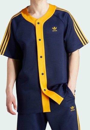 Person iført Adidas sportstøj i marineblåt og gult med korte ærmer, knaplukning foran og tre gule striber på ærmer og bukser.