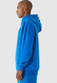 Felpa blu con una vestibilità rilassata, grande cappuccio, tasca frontale e polsini a costine. Presenta un branding minimale sul lato. Materiale morbido in cotone.
