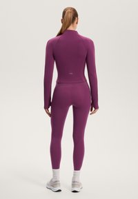 Těsně přiléhavé sportovní oblečení v tmavě fialové barvě, s vysokým límcem, dlouhými rukávy a sladěnými legínami. Elastický materiál s hladkou texturou.