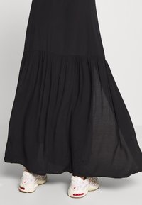 Jupe longue noire avec couture froncée, portée avec des baskets blanches sur un sol et un fond gris clair.