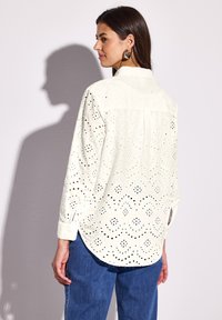 Valkoinen paita, jossa on pitsimäinen brodeeraus ja leikattuja kuvioita, pitkät hihat, pyöreä helma ja nappikaulus, yhdistetty sinisiin farkkuihin.