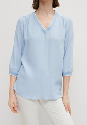 Vrouw draagt lichtblauw V-halsblouse met driekwart mouwen en witte broek tegen een effen achtergrond.