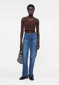 Γυναίκα που φοράει καφέ αμάνικο crop top, μπλε τζιν ψηλόμεσο με μεγάλες εμπρόσθιες τσέπες, μαύρα loafers και κρατάει ριγωτή πλεκτή τσάντα.