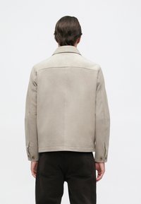 Giacca in suede beige con colletto strutturato, cuciture visibili e dettagli con bottoni a pressione. Design semplice con una texture liscia.