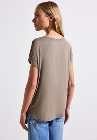 T-shirt marron clair à manches courtes avec une coupe décontractée, col rond et fentes sur les côtés. Texture douce, associé à un jeans bleu clair.
