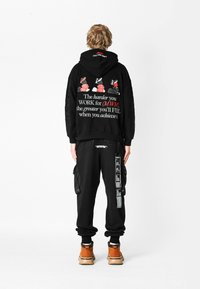 Persona vestida con sudadera con capucha negra y pantalones, con texto y gráficos en blanco y rojo en la parte de atrás, de pie contra un fondo blanco liso.