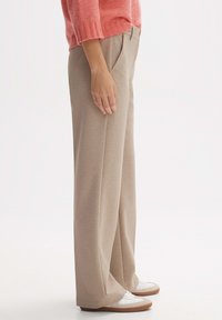 Beige weite Hosen aus weichem Stoff mit vorderen Falten, Seitentaschen und einer lockeren Passform. Kombiniert mit weißen Sneakers.