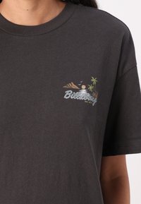 Maglietta nera in cotone con un piccolo ricamo grafico di montagne, palme e sole, con la scritta "Billabong" in lettere argentate.