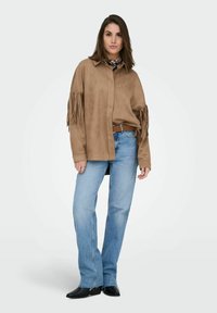 Brun ruskindsskjorte med frynsedetaljer på ærmerne, kombineret med lyseblå straight-leg jeans og sorte ankelstøvler. Tørklæde synligt ved halsen.