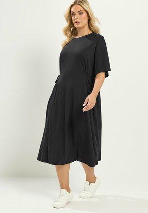 Frau trägt ein locker sitzendes schwarzes Midikleid mit kurzen Ärmeln und weißen Sneakers, steht vor einem schlichten hellen Hintergrund.