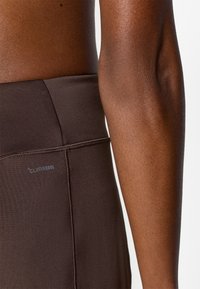 Καφέ κολάν ψηλόμεσο από αεριζόμενο ύφασμα, με λεία υφή και ετικέτα λογότυπου στο πλάι.