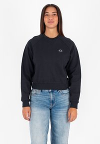Felpa nera corta con maniche raglan, polsini a costine e un piccolo logo bianco sul petto. Abbinata a jeans di denim azzurro chiaro.