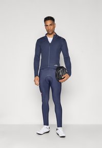 Marinefarbener Fahrradtrikot mit durchgehendem Reißverschluss, langen Ärmeln und Logo an der Taille, kombiniert mit eng anliegenden marineblauen Tights, hält einen schwarzen Helm und trägt weiße Schuhe.