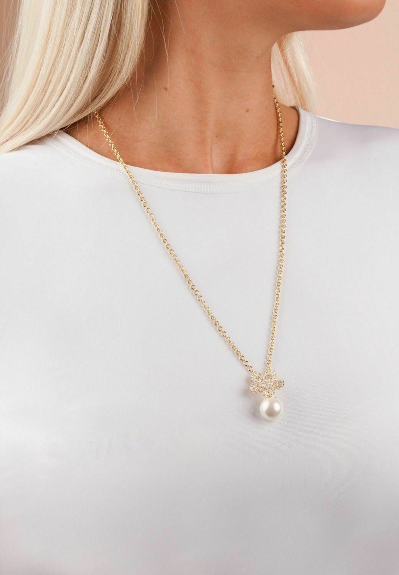 Guldkedja med en blommig design och ett pärlhänge. Kedjan har en mjuk textur mot en vit skjorta.