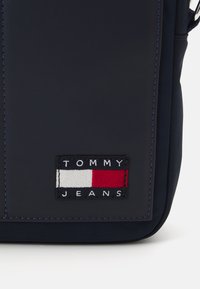 Bolso sintético de color azul marino con forma rectangular. Incluye un parche rectangular con "TOMMY JEANS" y un logotipo en rojo, blanco y azul.
