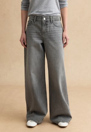 Persona che indossa jeans grigi a vita alta e a gamba larga con scarpe da ginnastica bianche, in piedi contro uno sfondo beige uniforme.