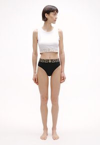 Λευκό cropped τοπ με χρυσό μοτίβο ελληνικού κλειδιού, σε συνδυασμό με ψηλόμεσο μαύρο σλιπ που διαθέτει παρόμοια ζώνη μέσης με χρυσές λεπτομέρειες.