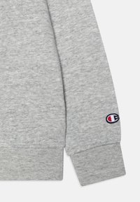 Manga de sudadera de algodón gris con puño acanalado; presenta un parche de logo bordado en azul, rojo y blanco en la zona de la muñeca.