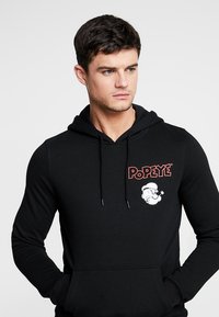 Fekete kapucnis pulóver, piros "Popeye" logóval és fehér rajzfilm tengerész grafikával a mellkasán. Kényelmes szabású.