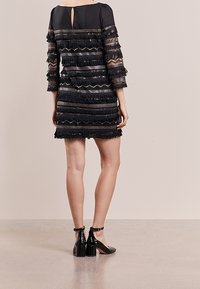 Robe noire à manches trois quarts, avec des détails en franges superposées et un dos en forme de goutte. Associée à des chaussures à talons blocs avec bride à la cheville noires.