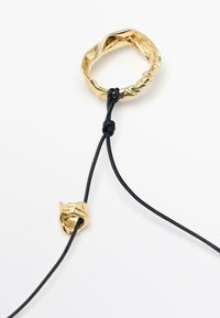 Anello in metallo dorato con un design ondulato, fissato a un cordino nero tramite un piccolo nodo. L'anello ha una superficie liscia e riflettente.