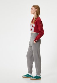 Afgeknotte kastanjebruine sweatshirt met het witte nummer 96, gecombineerd met grijze joggers met elastische tailleband en zakken, gedragen met teal sneakers.