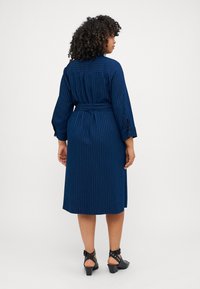 Abito blu navy con cintura, strisce verticali bianche, maniche lunghe, polsini bottonati e taglio dritto alla lunghezza del ginocchio. Tessuto strutturato.