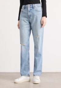 Lyseblå denimjeans med høy midje, rett passform, og to slitte lapper på framsiden. Kombinert med hvite sneakers.