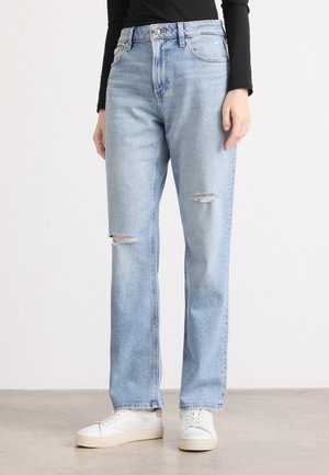 Jeans de denim azul claro con una cintura alta, corte recto y dos parches desgastados en la parte delantera. Combinados con zapatillas blancas.