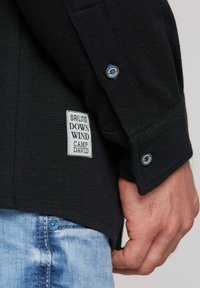 Camisa negra de manga larga texturizada con un parche de la marca y detalle de botones azules, combinada con jeans de mezclilla azul claro.