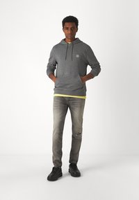 Grijze hoodie met voorste kangoeroezakken, trekkoordkap, gedragen over een gele shirt. Draagt een grijze spijkerbroek met een scheur, gecombineerd met zwarte sneakers.