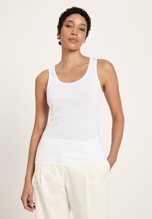 Woman with short curly hair wearing a white sleeveless tank top and cream-colored pants, standing with one hand in pocket.