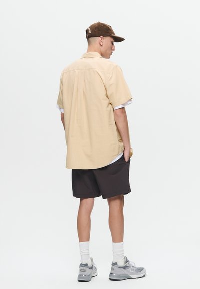 Chemise jaune clair à manches courtes avec une coupe décontractée, associée à un short noir. Des baskets grises et une casquette marron complètent la tenue.