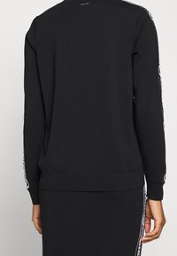 Czarny sweatshirt z prążkowanym dekoltem w serek i mankietami, z kontrastowymi białymi nadrukami na rękawach i u dołu. Gładka tekstura.