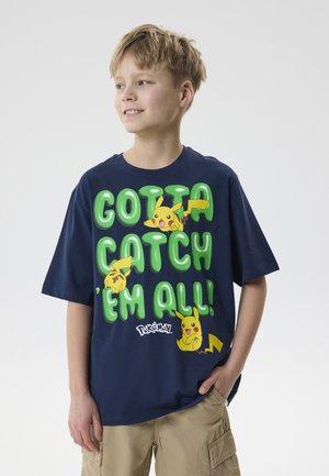 Αγόρι που φοράει ναυτικό μπλε μπλουζάκι με πράσινο κείμενο "Gotta Catch 'Em All!" και τρία γραφικά Pikachu, συνδυασμένο με μπεζ cargo σόρτς.