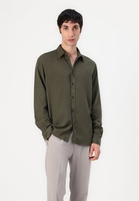 Camisa de manga larga verde oliva con cierre de botones y un corte holgado. El material parece suave con una textura sutil. Se lleva con pantalones beige claros.